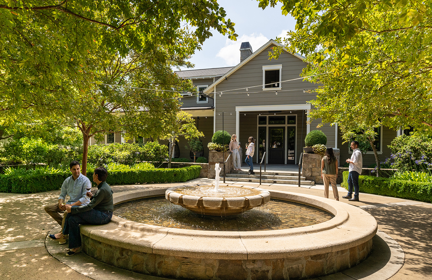 Main building of Trinchero Napa Valley with a fountain in the front surrounded by lush green trees with people enjoying glasses of wine in the sunshine