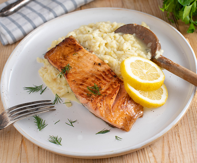 A white plate with a seared salmon fillet topped with dill and slices of lemon on a bed of creamy risotto being served with a wooden spoon