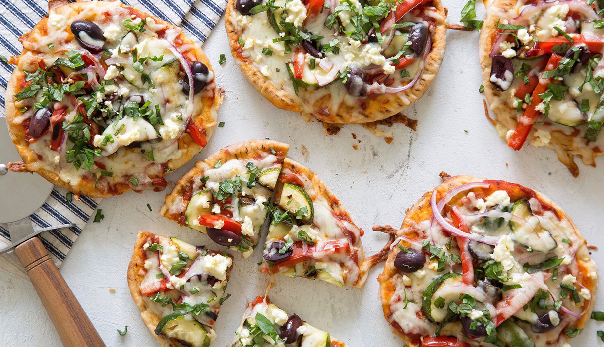 Five mini pizzas topped with veggies and cheese, one cut into four slices with a pizza cutter and dishtowel nearby.