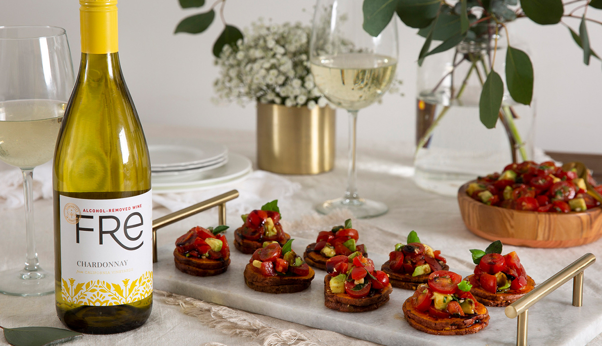 A platter of sweet potato crostini with avocado bruschetta topping on a dining table next to a bottle and glasses of FRE alcohol-removed chardonnay and vases of small white flowers and greenery in the background.