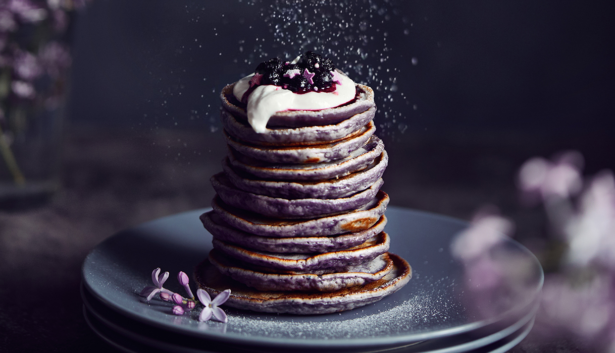 purple pancakes with whipped cream and blueberries on top being dusted with powdered sugar.