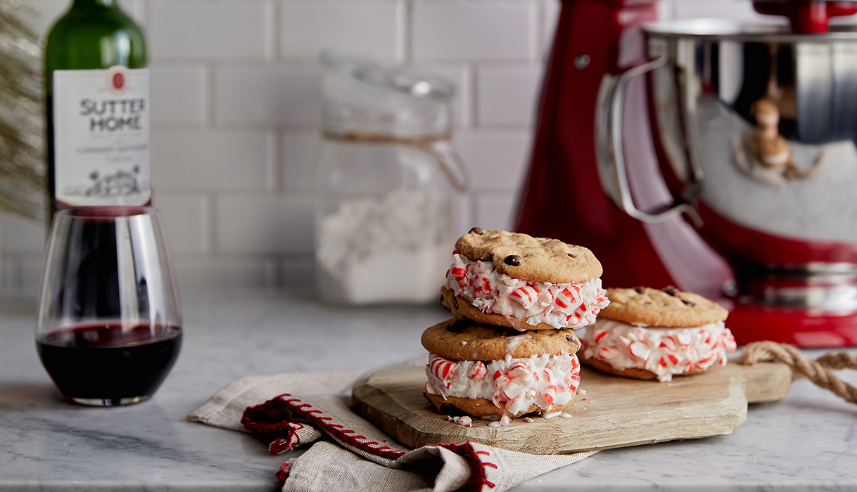 Three peppermint icecream cookie sandwiches stacked on a kitchen counter with a bottle and glass of Sutter Home cabernet sauvignon to the left and a red stand mixer in the background.