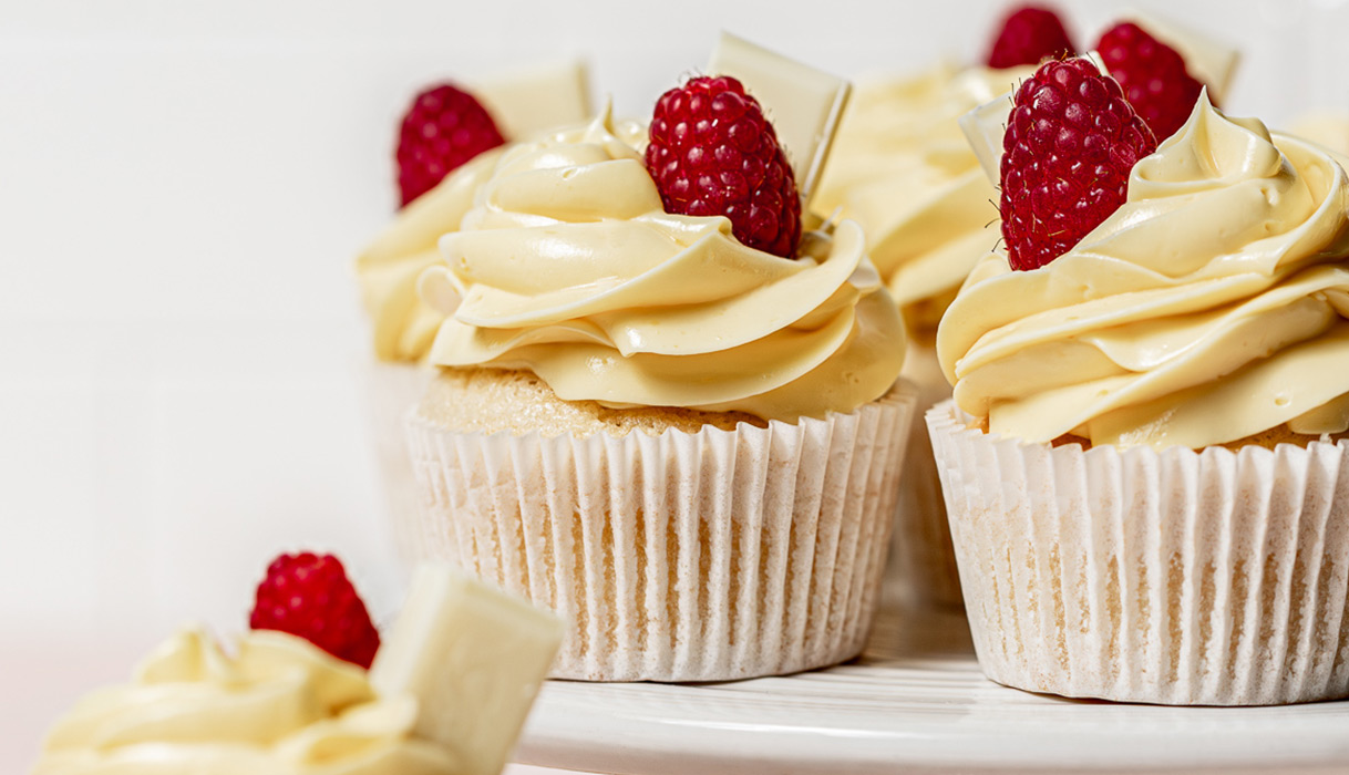a tray of white chocolate raspberry cupcakes garnished chunks of white chocolate and fresh, bright red raspberries. Learn how to make this dessert recipe from One Stop Wine Shop.