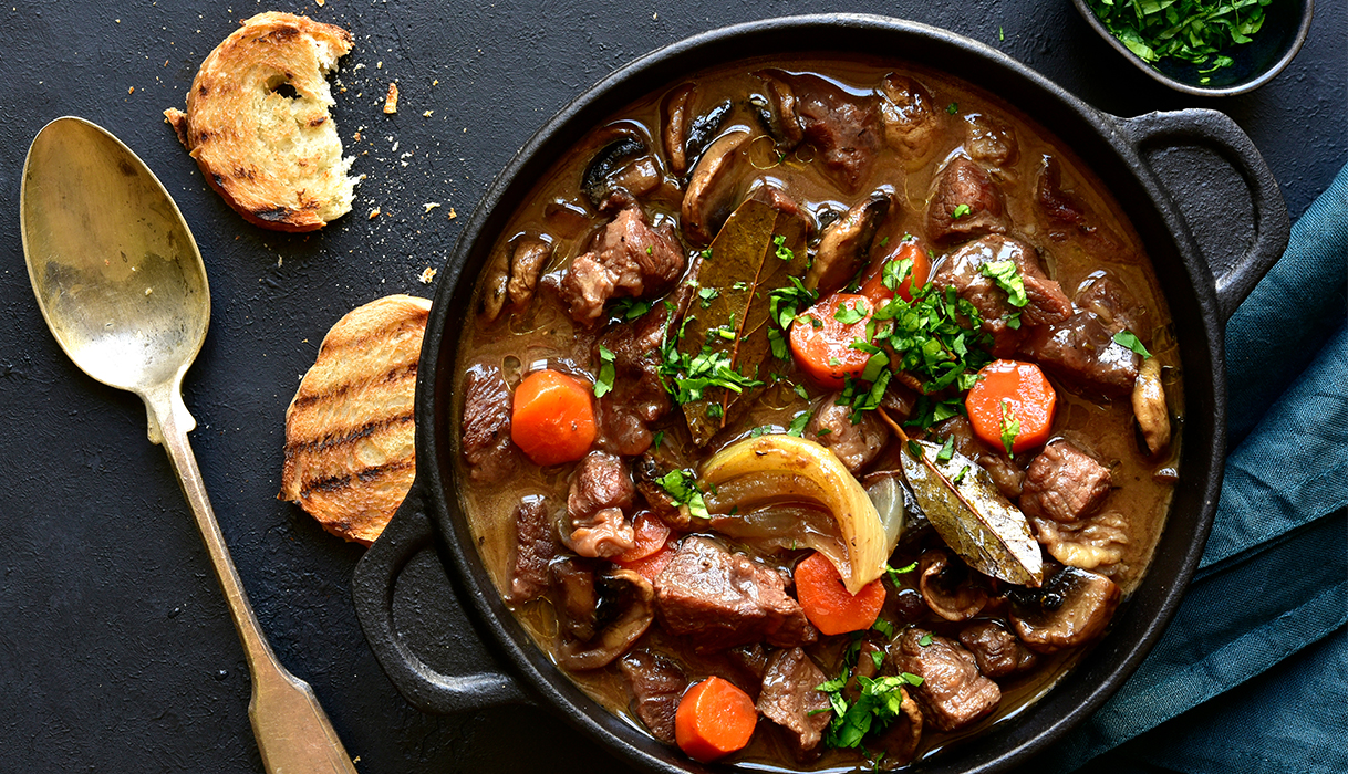 A hearty bowl of boeuf bourguignon on a table next to a gold spoon and some toasted pieces of bread.