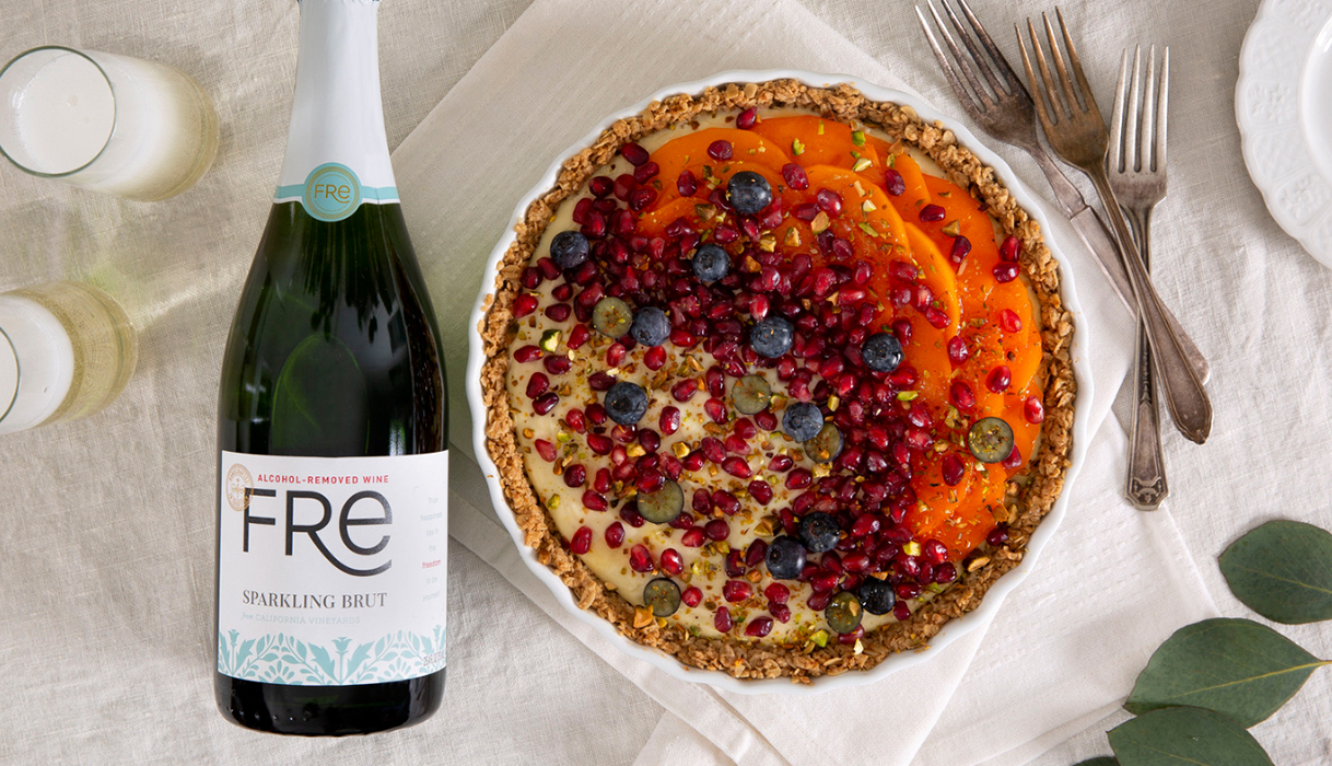 bottle of FRE alcohol-removed sparkling brut next to a honey greek yogurt tart decorated with fruit sitting on a white tablecloth with leaves and flowers in the background
