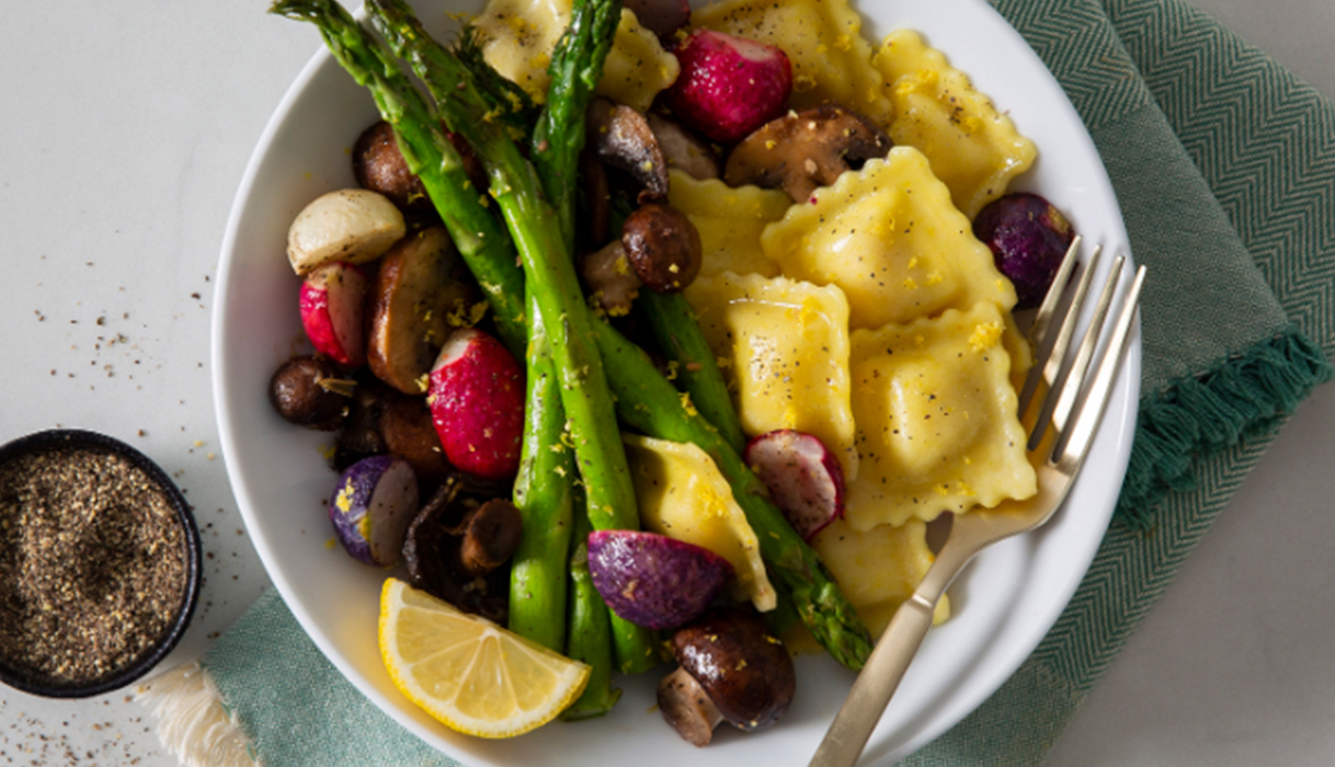 A while bowl of ravioli and roasted mushrooms, asparagus, and radishes with a fork and a light blue napkin, and a bowl of freshly ground pepper next to it.