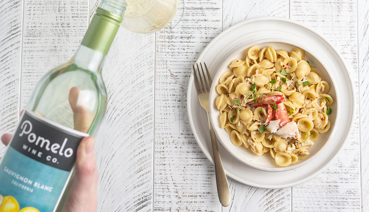 hand pouring a bottle of pomelo sauvignon blanc next to a bowl of spicy crab pasta on a white wood surface.