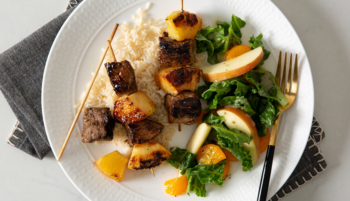 a plate of steak and pineapple kebabs on a bed of rice next to an apple and kale salad.