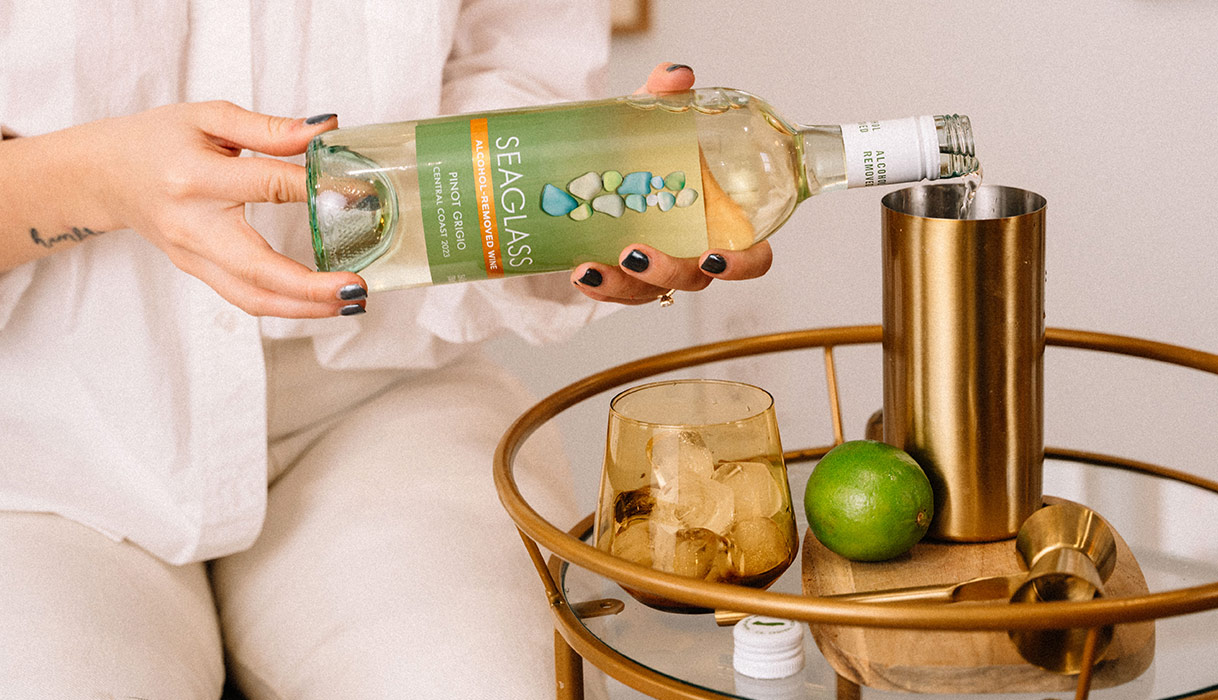 woman in a white button-up shirt pouring a bottle of SEAGLASS alcohol-removed pinot grigio into a cocktail shaker next to a glass full of ice and a lime.