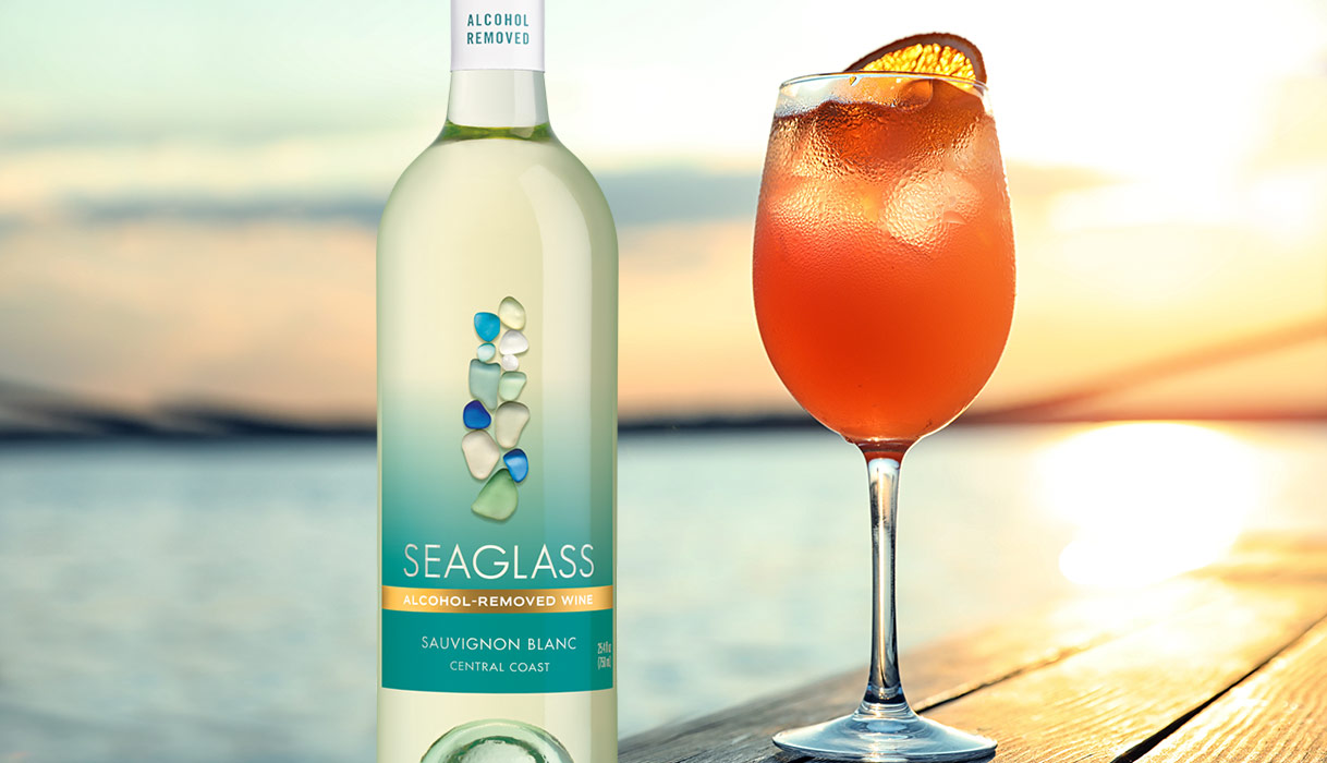 a sunset spritz mocktail in a glass next to a bottle of SEAGLASS alcohol-removed sauvignon blanc with the sun setting over the ocean in the background.