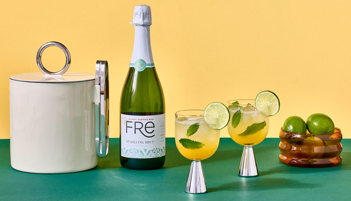 Glasses of FRE mango mojitos garnished with lime circles next to a bowl of limes, and ice bucket, and a bottle of FRE alcohol-removed sparkling brut. Get this summer mocktail recipe and more.