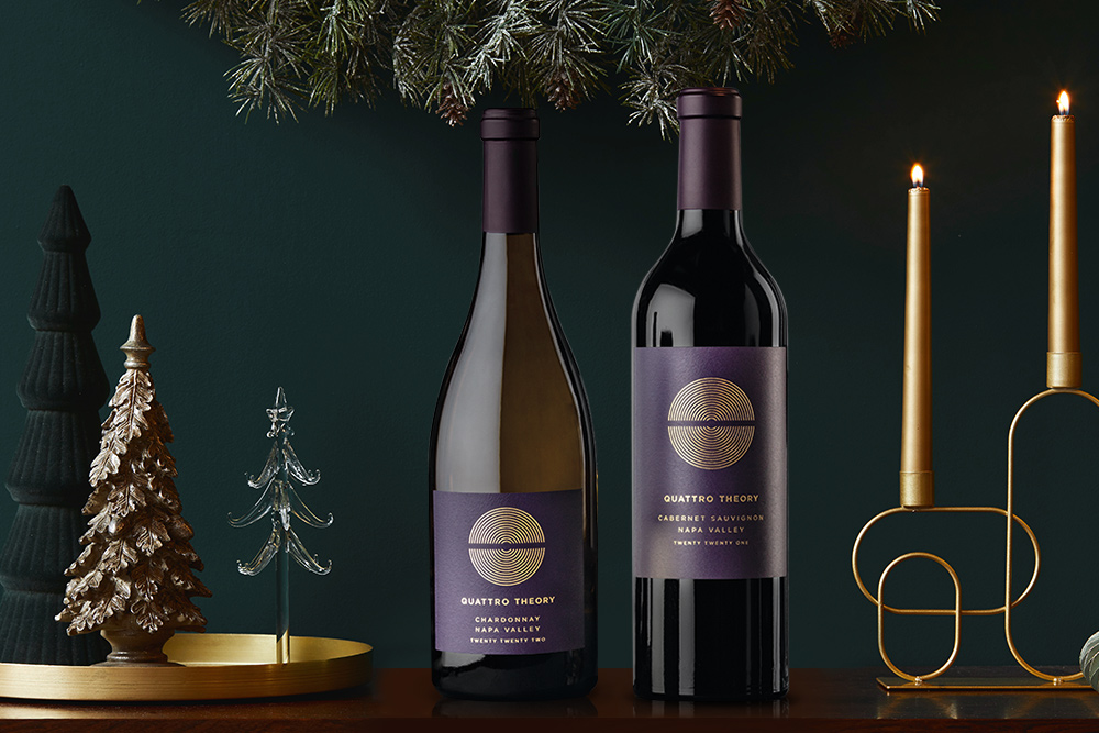 A bottle of Quattro Theory chardonnay and cabernet sauvignon on a wooden table with festive green and gold decorations and a green wall in the background. Shop this and more custom wine gift sets from One Stop Wine Shop.