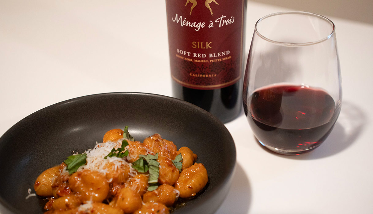 A bowl of gnocchi topped with fresh basil and parmesan cheese next to a bottle and glass of Ménage à Trois silk red blend. Get this recipe and more at One Stop Wine Shop!