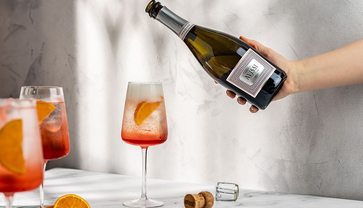 Bubbly pink glasses of Negroni Sbagliato Spritzes garnished with orange slices next to a bottle of Avissi Prosecco. Get this cocktail recipe and more from One Stop Wine Shop.